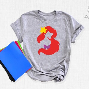 May include: A gray t-shirt with a red silhouette of Ariel, a Disney princess, with a yellow flower in her hair. The silhouette is in a simple, minimalist style.