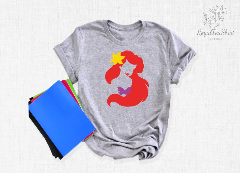 May include: A gray t-shirt with a red silhouette of Ariel, a Disney princess, with a yellow flower in her hair. The silhouette is in a simple, minimalist style.