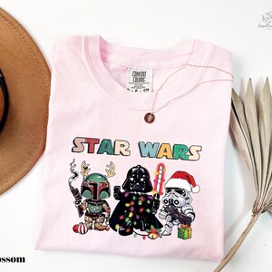 A pink t-shirt with a Star Wars-themed graphic design featuring three characters: a green elf, a black knight, and a white stormtrooper, along with Christmas decorations.