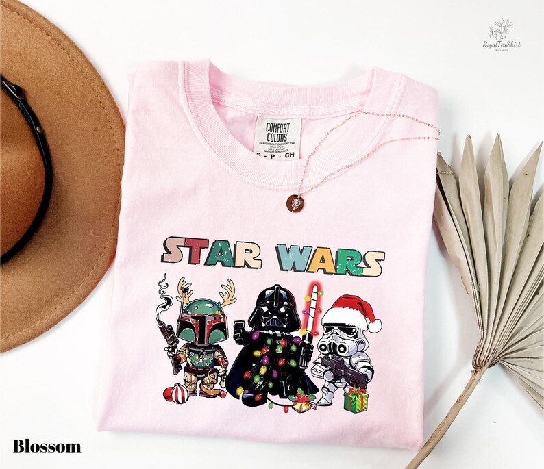 A pink t-shirt with a Star Wars-themed graphic design featuring three characters: a green elf, a black knight, and a white stormtrooper, along with Christmas decorations.
