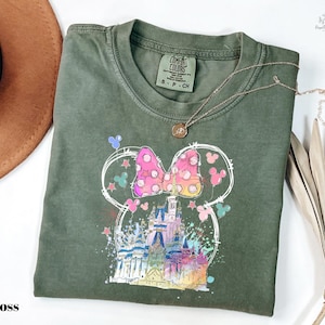 a green t-shirt with a colorful Mickey Mouse design, including a castle and various Disney-themed elements. The t-shirt is folded and placed on a white surface, with a brown hat and some dried leaves nearby.