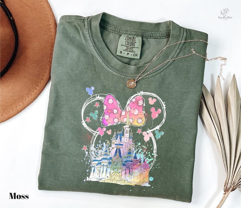a green t-shirt with a colorful Mickey Mouse design, including a castle and various Disney-themed elements. The t-shirt is folded and placed on a white surface, with a brown hat and some dried leaves nearby.