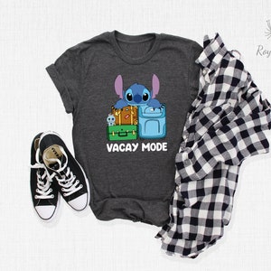 May include: A dark heather grey t-shirt with a cartoon Stitch character from Lilo & Stitch. Stitch is holding onto a stack of luggage with the text "Vacay Mode" printed below.