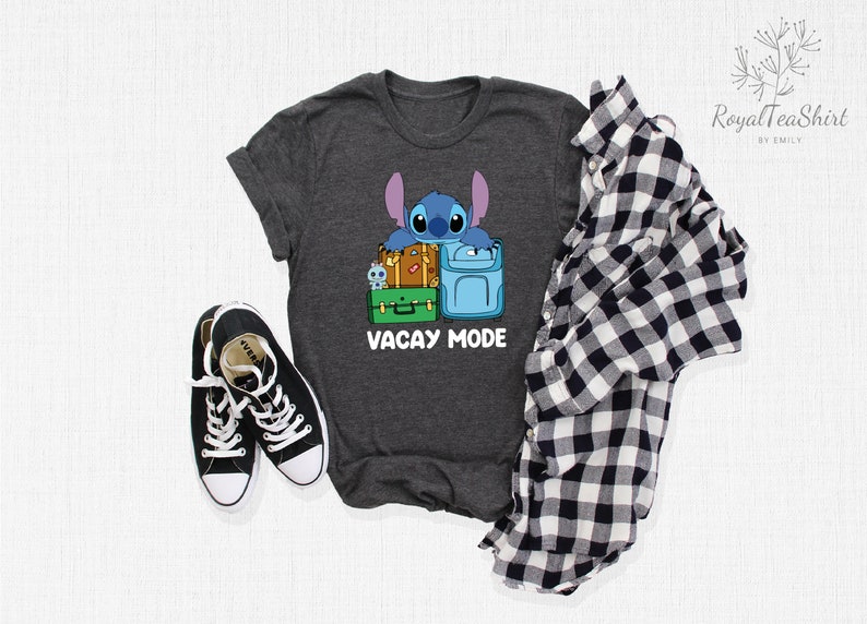 May include: A dark heather grey t-shirt with a cartoon Stitch character from Lilo & Stitch. Stitch is holding onto a stack of luggage with the text "Vacay Mode" printed below.