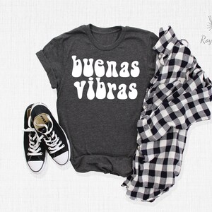 May include: A dark gray t-shirt with the words "buenas vibras" in white, retro-style lettering. The shirt is laid flat on a white surface with a black and white plaid shirt and black sneakers.