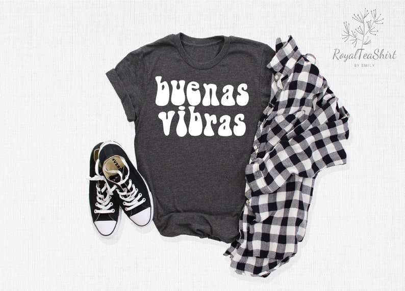 May include: A dark gray t-shirt with the words "buenas vibras" in white, retro-style lettering. The shirt is laid flat on a white surface with a black and white plaid shirt and black sneakers.