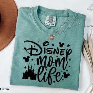 A light green t-shirt with a Disney-themed design, including a castle silhouette and stars, along with the text "Disney mom life" printed on it. The t-shirt is folded and placed on a white surface, with a brown hat and some dried leaves nearby.