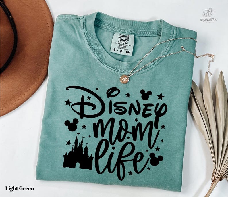 A light green t-shirt with a Disney-themed design, including a castle silhouette and stars, along with the text "Disney mom life" printed on it. The t-shirt is folded and placed on a white surface, with a brown hat and some dried leaves nearby.