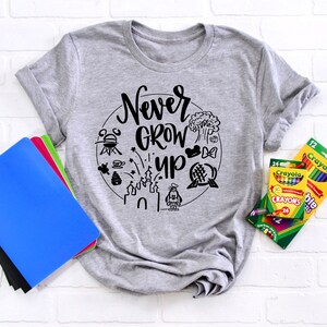May include: Gray t-shirt with a black graphic that says "Never Grow Up" surrounded by Disney-themed icons. The shirt is laying on a white brick background with a stack of notebooks and a box of crayons.