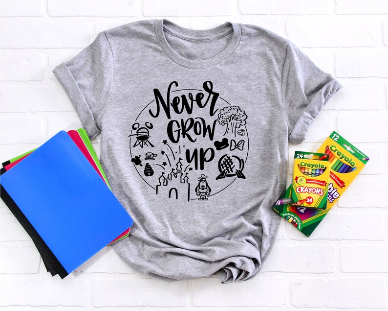 May include: Gray t-shirt with a black graphic that says "Never Grow Up" surrounded by Disney-themed icons. The shirt is laying on a white brick background with a stack of notebooks and a box of crayons.
