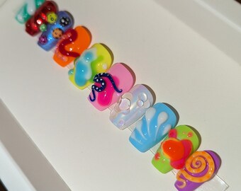 May include: A set of ten colorful fake nails with various designs, including swirls, seashells, and snakes. The nails are arranged in a row on a white surface.