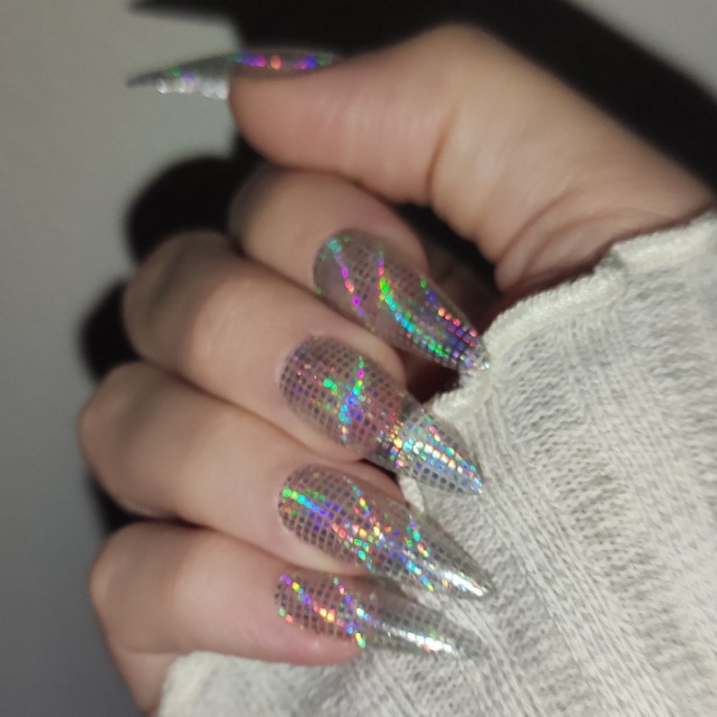 May include: Close-up of a hand with long, pointed nails. Each nail is covered in a holographic, iridescent glitter polish.