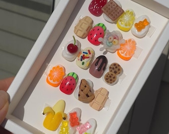 Puede incluir: Una colección de diseños decorativos para uñas, que incluyen fresas en miniatura, barras de chocolate y ositos de goma. El arte de uñas está dispuesto en una caja blanca, mostrando varios diseños coloridos con temática de comida.