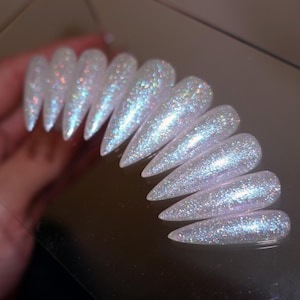 May include: A set of stiletto-shaped, iridescent press-on nails. The nails are a pearly white color and covered in glitter, reflecting a rainbow of colors. The nails are arranged in a row, showcasing their pointed shape and shimmering finish.