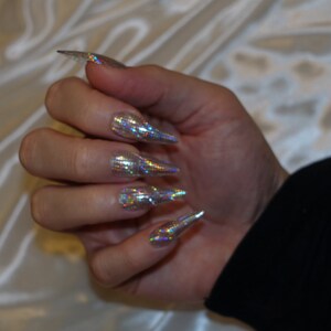 May include: Close-up of a hand with long, pointed nails. The nails are covered in a silver glitter polish with a holographic finish.