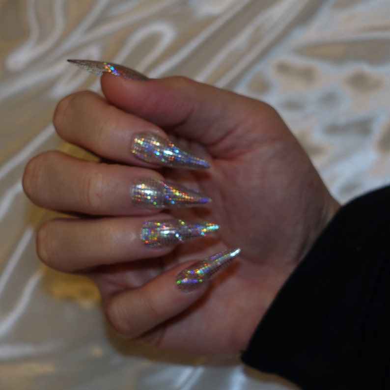 May include: Close-up of a hand with long, pointed nails. The nails are covered in a silver glitter polish with a holographic finish.