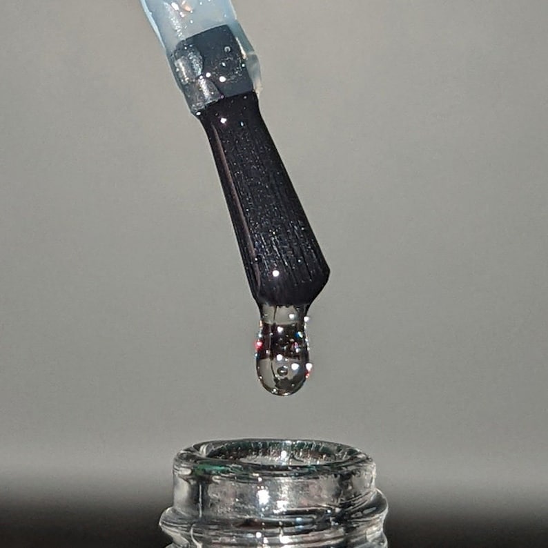 May include: Close-up of a black nail polish brush with a single drop of clear polish suspended above a bottle. The polish and bottle have glittery accents, reflecting light. The image is set against a neutral gray background.