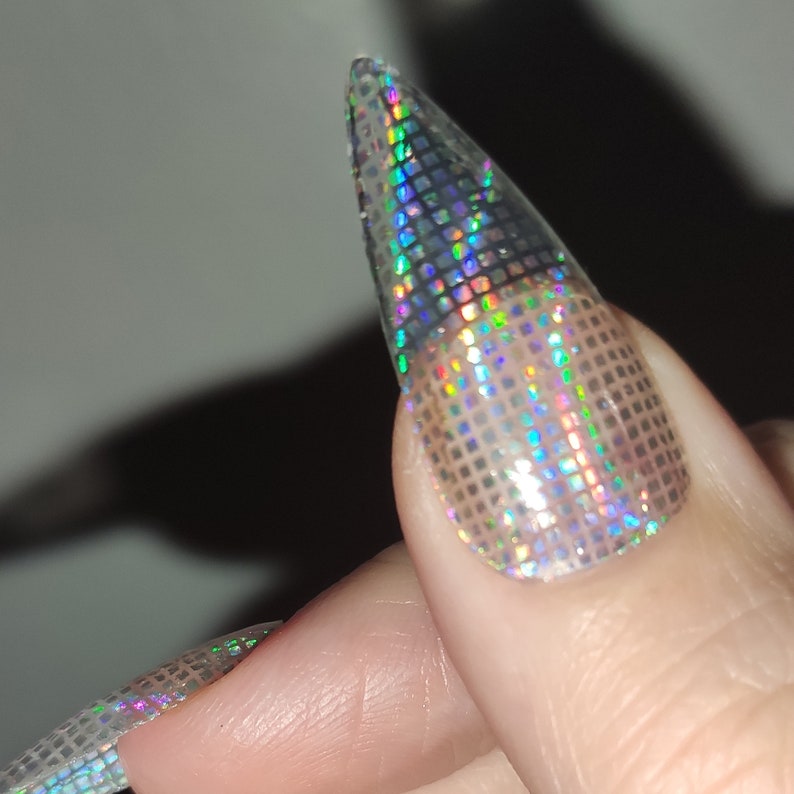 May include: Close-up of a long, pointed, clear acrylic nail with a holographic, iridescent, square-shaped glitter design.