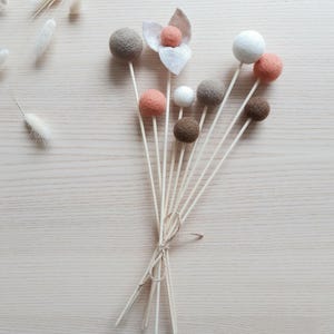 Pode incluir: Um buquê de flores decorativas de feltro em tons de marrom, pêssego e branco. As bolas de feltro são presas a pinos de madeira e amarradas com barbante. Uma flor de feltro com uma folha também está incluída.