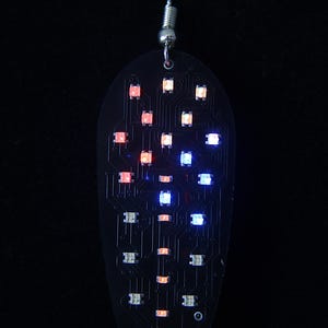 May include: A single, teardrop-shaped earring with a black circuit board design. The earring features an array of small, illuminated LED lights in red, white, and blue. The earring hangs from a silver-colored hook.