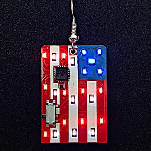 May include: A unique, patriotic earring featuring a circuit board design in the shape of the American flag. The red and white stripes and blue corner are illuminated with small LED lights. The earring hangs from a silver-colored hook.