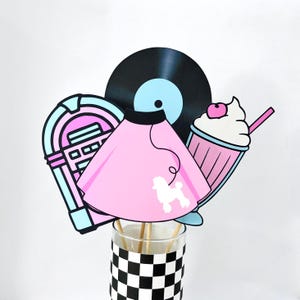 May include: A collection of 1950s-themed cutouts, including a pink poodle skirt, a jukebox, a vinyl record, and a milkshake. The cutouts are arranged in a glass with a black and white checkered pattern.