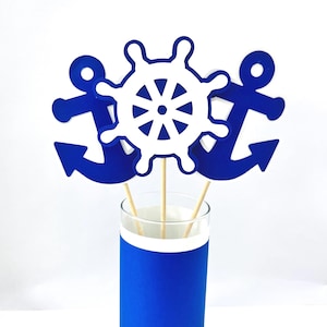 May include: Nautical-themed decorations featuring a white ship's wheel and blue anchors. The decorations are attached to wooden sticks and placed in a glass with a blue base. The background is white.