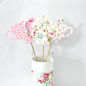 May include: Three decorative tea party props in a glass vase. The props are shaped like a teacup and teapot, with floral patterns in pink, white, and blue. The vase has a white band and a floral pattern at the bottom.