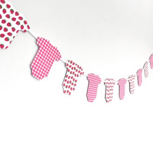 May include: A decorative banner featuring baby onesie cutouts. The onesies have various patterns, including pink gingham and white with red strawberries. The banner is strung on a white string and is against a white background.