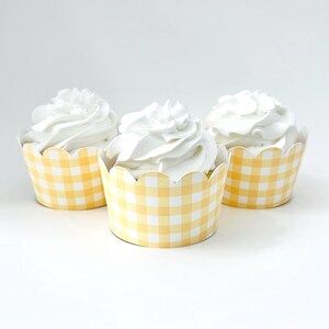 May include: Three cupcakes topped with white swirled frosting, each encased in a yellow and white gingham patterned paper wrapper with a scalloped top edge. The cupcakes are arranged in a triangular formation against a plain white background.