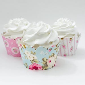 May include: Three cupcakes with white frosting in decorative paper liners. The liners feature floral patterns in pink, blue, and white. The cupcakes are arranged on a white surface.