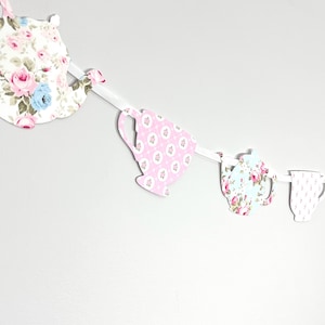 May include: A decorative banner featuring cut-out shapes of teacups and teapots. The shapes are adorned with floral patterns in pink, blue, and white, strung together with a white ribbon. The banner is designed for a tea party or vintage-themed event.