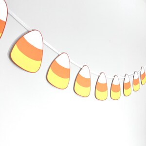 May include: A string of candy corn decorations, perfect for Halloween. Each candy corn is white, orange, and yellow, strung together on a white string. The candy corn shapes are in various sizes, creating a festive look.