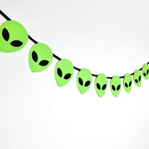 May include: A decorative garland featuring a series of bright lime green alien head cutouts strung along a thin black cord. Each alien head shape displays two large, almond-shaped black eyes, creating a repeating pattern across the length.