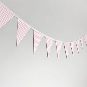 May include: A string of pink and white gingham pattern pennant flags. The triangular flags are strung together on a white ribbon. The flags are hanging against a plain, light gray background.