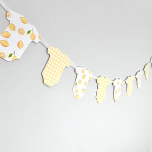 May include: A baby shower banner featuring onesie cutouts. The onesies are decorated with lemon and gingham patterns in yellow and white. The banner is strung on a white ribbon and hangs against a plain white wall.