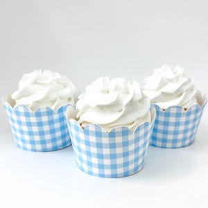 May include: Three cupcakes with white frosting in blue and white gingham patterned cupcake liners. The liners have a scalloped edge. The cupcakes are arranged on a white surface.