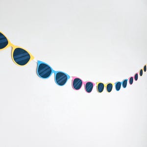 May include: A decorative garland featuring a series of paper sunglasses in various bright colors including yellow, blue, and pink. Each pair of sunglasses has dark blue lenses with white diagonal lines, connected by a thin white string in a row.