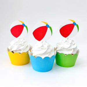 May include: Three cupcakes topped with white frosting and round paper beach ball decorations featuring red, white, blue, and green segments. The cupcakes are held in scalloped paper wrappers in solid yellow, bright blue, and lime green colors.