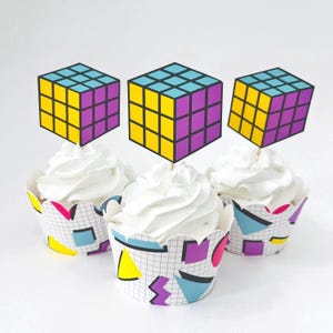 May include: Three cupcakes topped with paper cube puzzle decorations featuring yellow, blue, and purple grids. The cupcake wrappers are white with a black grid pattern and colorful geometric shapes in yellow, blue, and purple accents.