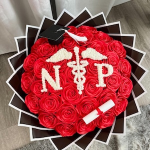 May include: A bouquet of red roses arranged in a circular shape, with the letters "N P" and a medical symbol in the center. A small graduation cap and a rolled diploma are included. The bouquet is wrapped in black and white patterned paper.