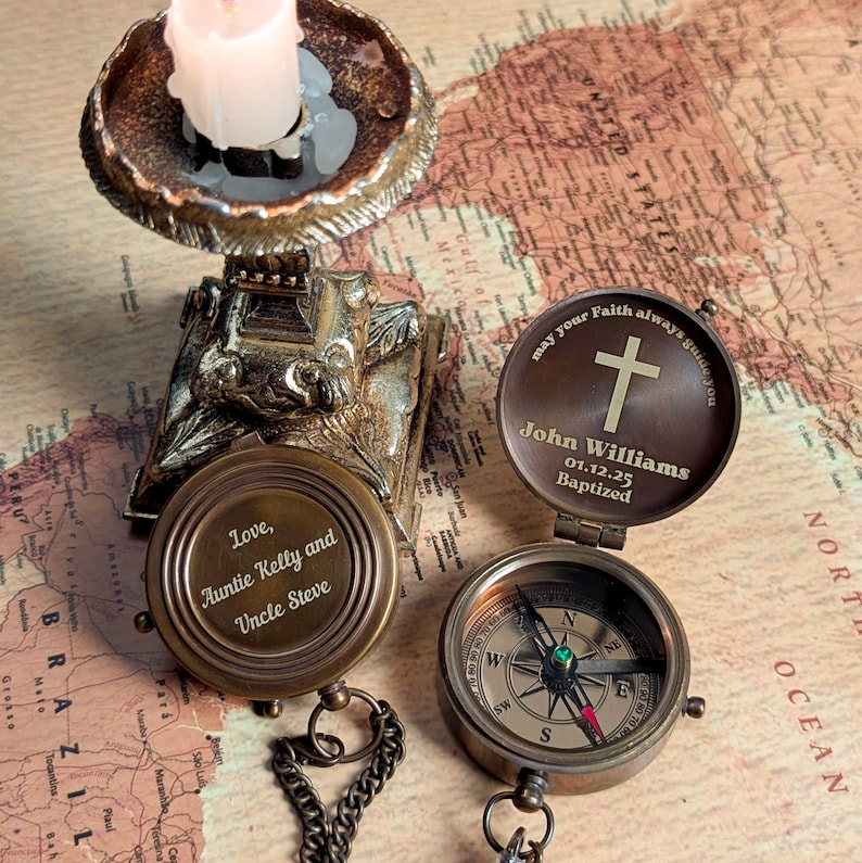 May include: A brass compass with a chain and a lid that opens to reveal a personalized inscription. The inscription reads "May your faith always guide you, John Williams, 01.12.25 Baptized". The compass is on a map background.