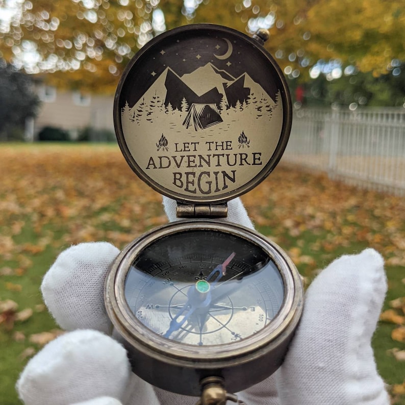 May include: A brass compass with a black and white design featuring mountains, a tent, and the text "Let the Adventure Begin". The compass is open and the needle is pointing north.