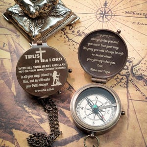 May include: A brass compass with a chain, featuring a quote about trusting in the Lord. The compass is open, revealing a compass face with a green jewel in the center. The quote reads: "TRUST in the LORD WITH ALL YOUR HEART AND LEAN NOT ON YOUR OWN UNDERSTANDING: in all your ways submit to Him, and He will make your Paths straight Proverbs 3:5-6". The other side of the compass has a message that reads: "May your faun always guide you and you never lose your way. We pray you will always be safe no matter where your journey takes you. Love, Nana and Papa."