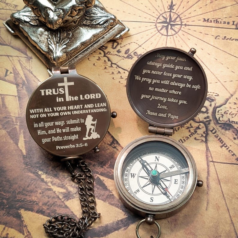 May include: A brass compass with a chain, featuring a quote about trusting in the Lord. The compass is open, revealing a compass face with a green jewel in the center. The quote reads: "TRUST in the LORD WITH ALL YOUR HEART AND LEAN NOT ON YOUR OWN UNDERSTANDING: in all your ways submit to Him, and He will make your Paths straight Proverbs 3:5-6". The other side of the compass has a message that reads: "May your faun always guide you and you never lose your way. We pray you will always be safe no matter where your journey takes you. Love, Nana and Papa."