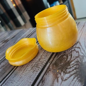 Puede incluir: Un recipiente de plástico amarillo con tapa. El recipiente es redondo y tiene un acabado liso y brillante. La tapa también es redonda y tiene un borde ondulado.