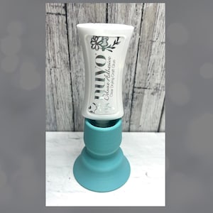 May include: A white tube of Nuvo Deluxe Adhesive, a clear-drying craft glue, sits atop a turquoise-colored, decorative stand. The tube has black floral designs and text. The stand has a flared base and a cylindrical top.