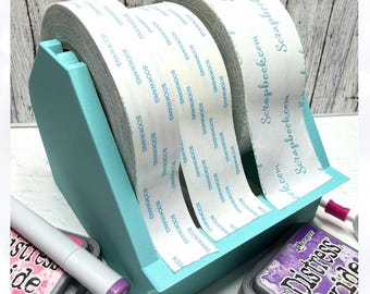 May include: A teal tape dispenser holding rolls of tape labelled "SOOKWANG" and "Scrapbook.com". A purple ink pad and a marker are visible in the foreground. The dispenser is designed for crafting.