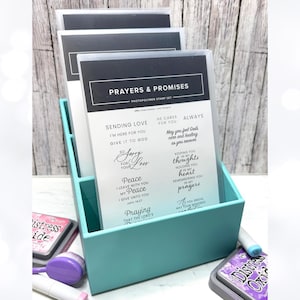 May include: A teal organizer holds several clear stamp sets labeled "Prayers & Promises." The stamps feature various inspirational phrases and sentiments. Distress Oxide ink pads and blending tools are visible in the foreground.