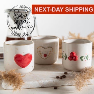 Puede incluir: Tres tazas de cerámica blanca con ribete dorado y diferentes diseños navideños. Las tazas presentan un corazón rojo, un reno y una rama de acebo con bayas rojas.